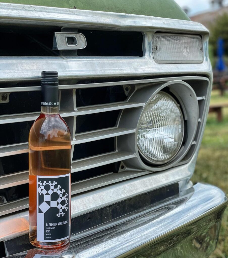 A bottle of Blenheim Vineyards Rosé Wine 2020 is propped against the grille of a vintage green pickup truck. The rosé’s pale pink hue contrasts with the truck’s silver and green tones, evoking a rustic, laid-back atmosphere.