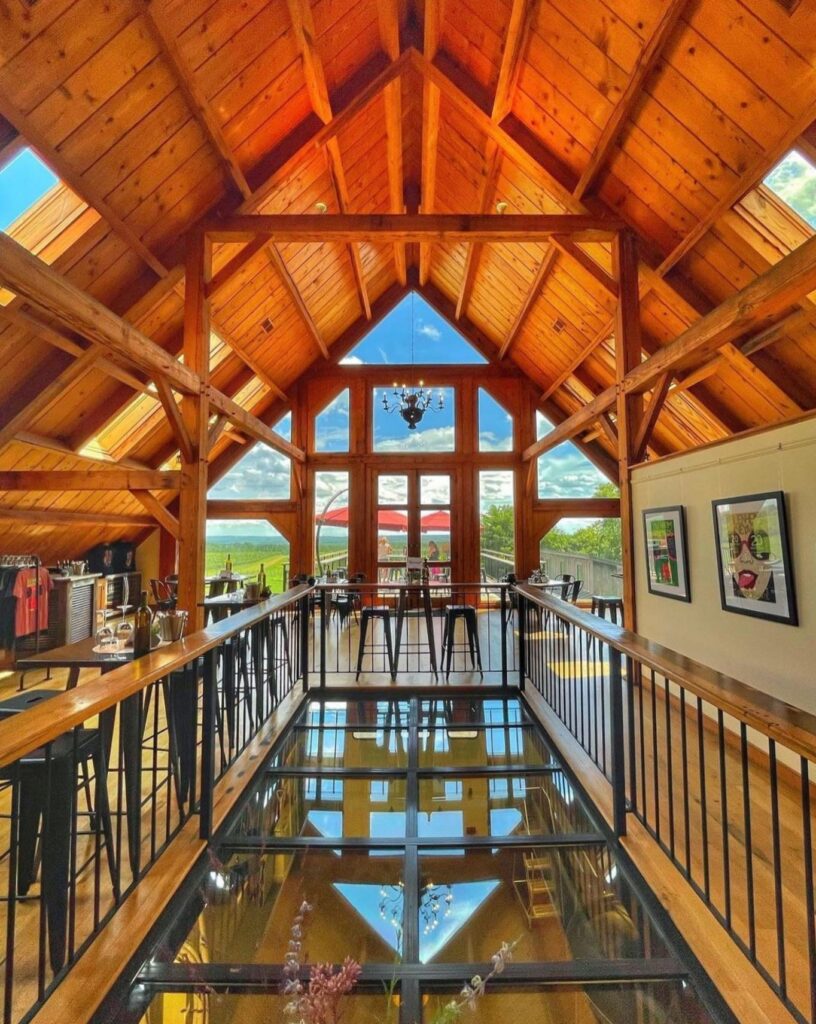A warm, wooden tasting room with high vaulted ceilings and large windows showcasing a vineyard view. The interior features a glass floor section, black metal railings, modern stools, framed artwork, and retail displays.