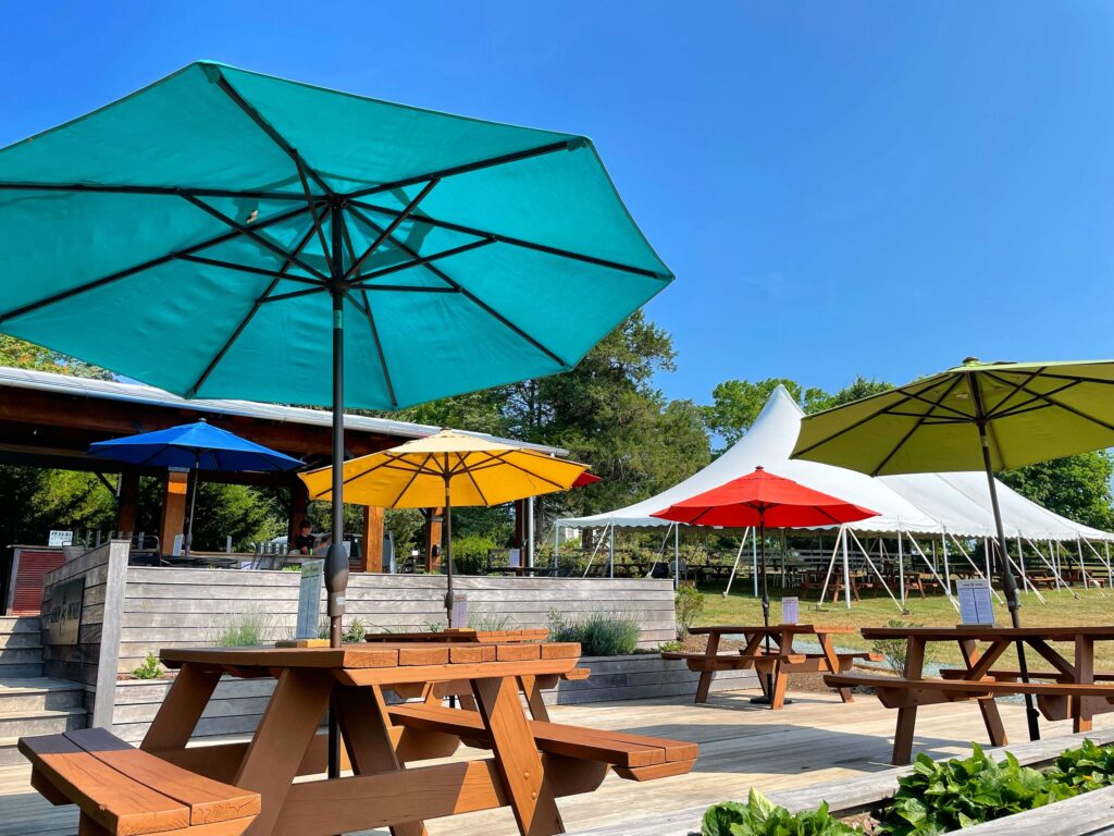 Wooden picnic tables with brightly colored umbrellas—teal, yellow, red, green, and blue—set up on a sunny outdoor deck next to a white event tent and a shaded pavilion surrounded by trees and clear blue sky.