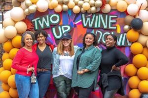 Five women smile and pose in front of a vibrant balloon arch and backdrop that reads "TWO UP WINE DOWN" at an outdoor wine event. The backdrop features bold, colorful patterns and is framed by balloons in shades of gold, cream, orange, and black. The women are casually dressed and holding wine bottles or glasses, enjoying the festive atmosphere of the gathering.