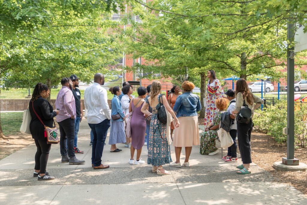 A diverse group of people gathers outdoors under a canopy of green trees, listening attentively to a woman in a colorful dress who is addressing the group. The setting is a shaded urban park with visible buildings and cars in the background.