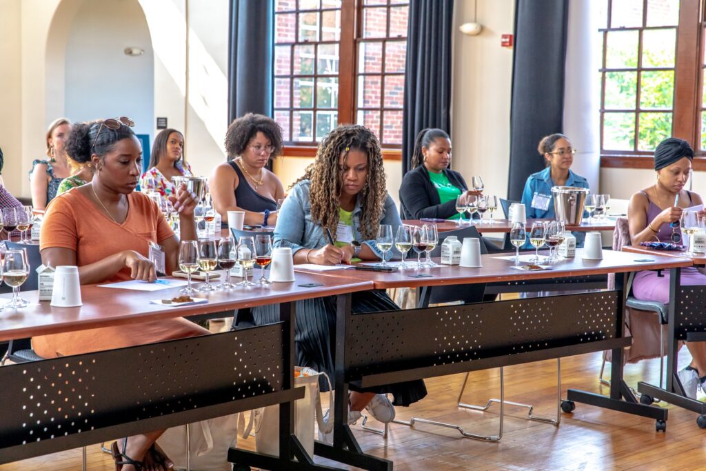 A diverse group of participants at an Oenoverse event sit attentively in a sunlit room, engaging in a guided wine tasting. Each person has multiple glasses of wine, tasting notes, and educational materials in front of them. The mood is focused and reflective, highlighting a learning atmosphere centered on wine appreciation and community.