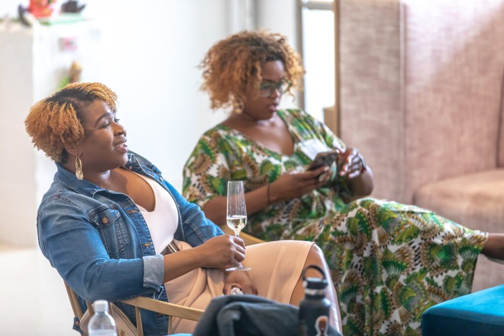 Two women sit comfortably indoors, enjoying a moment of relaxation. The woman in the foreground wears a denim jacket and holds a flute of sparkling wine, smiling as she listens. The woman in the background, dressed in a colorful green patterned dress, focuses on her phone. The atmosphere feels casual and inviting.