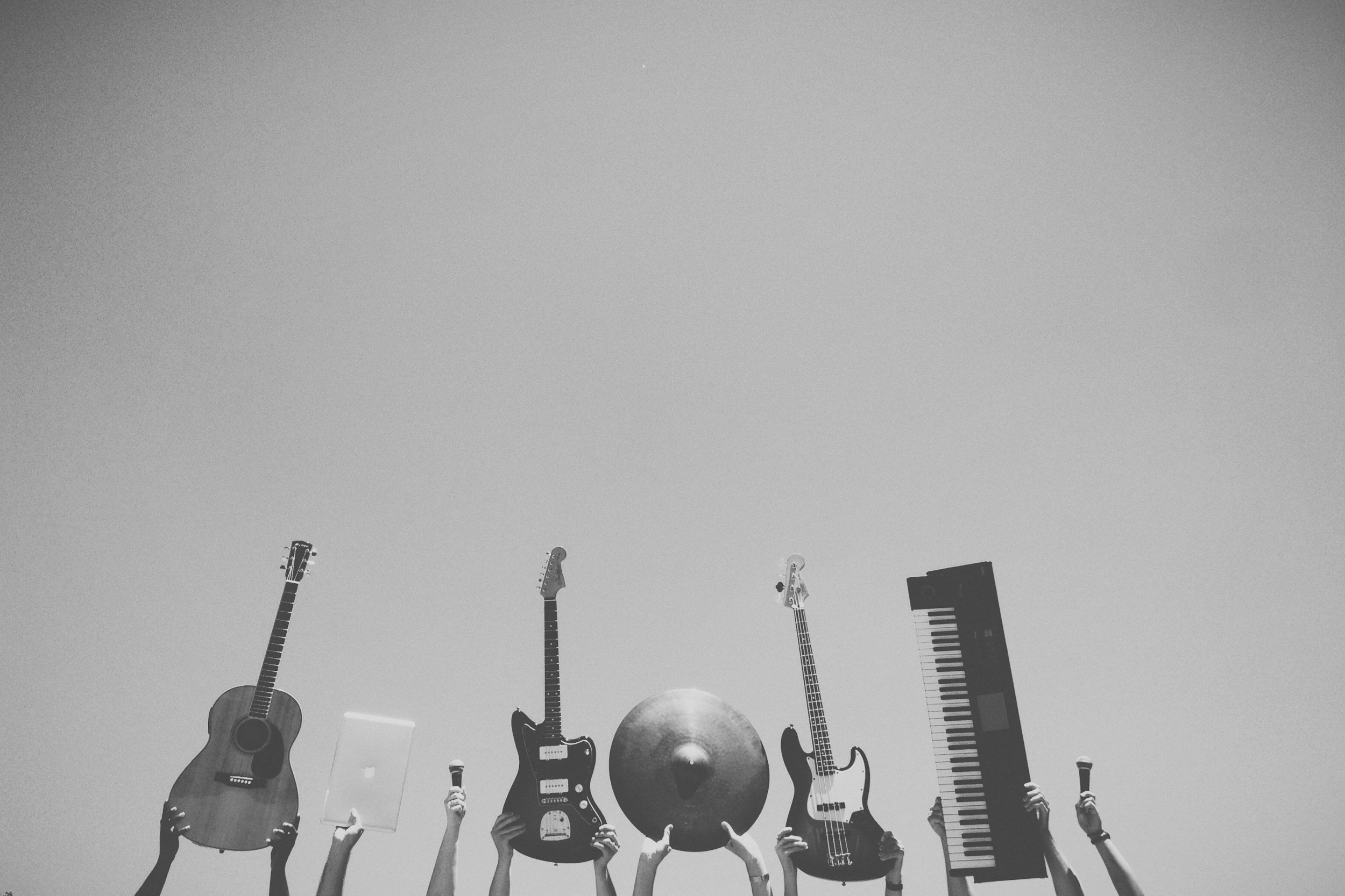 Black and white photo, with hands holding instruments and technology at the bottom of the page. From left to right, the hands hold an acoustic guitar, a macbook, a microphone, an electric guitar, a cymbal, an electric guitar, a piano keyboard, and a microphone.