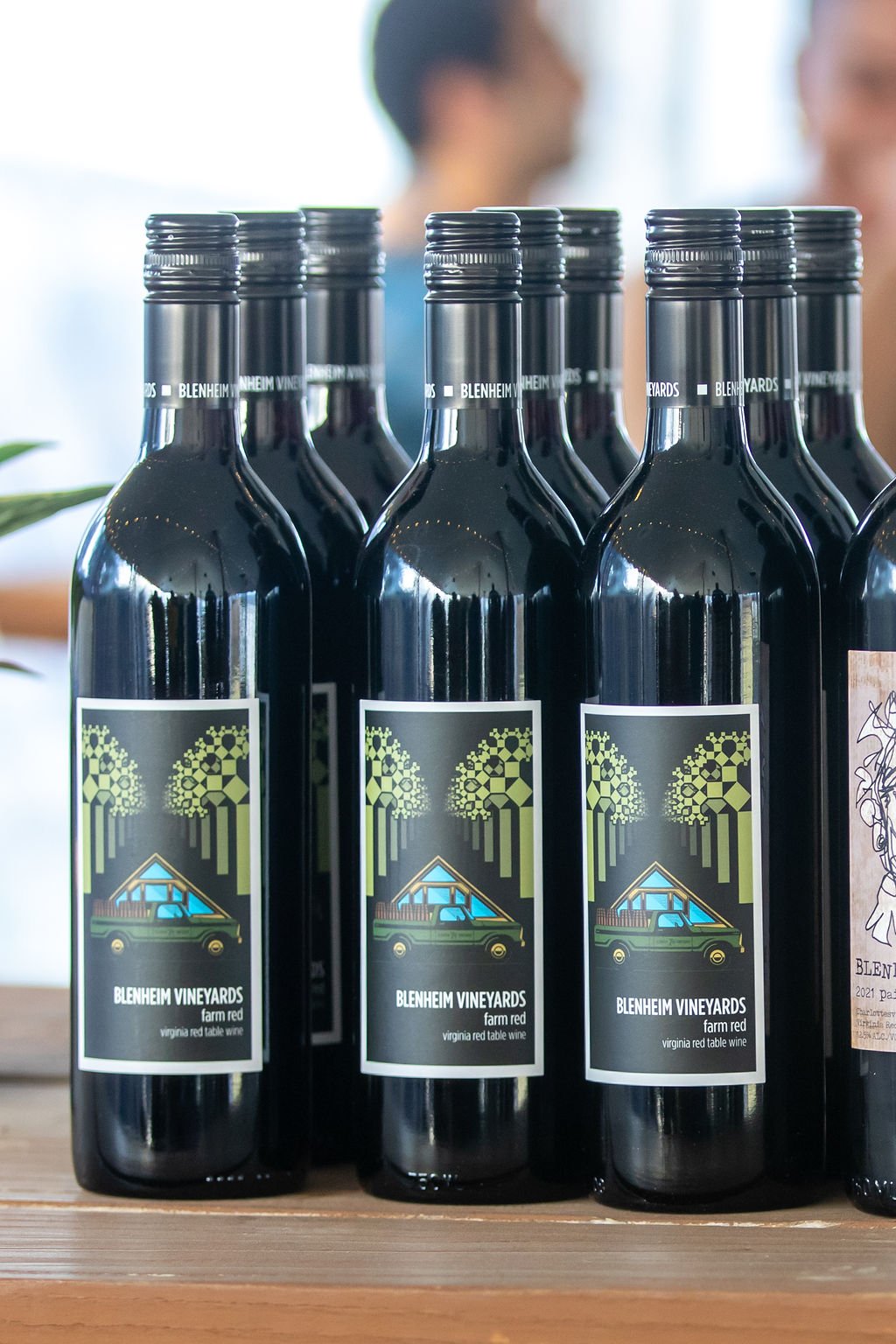 A group of Blenheim Vineyards "Farm Red" wine bottles arranged on a wooden surface. The label features a stylized illustration of a vehicle with a house-like structure on top, set against a background of geometric trees with checkerboard patterns. The text reads "BLENHEIM VINEYARDS farm red, virginia red table wine." The black screw-top bottles are positioned closely together, with a blurred background of people and natural light.
