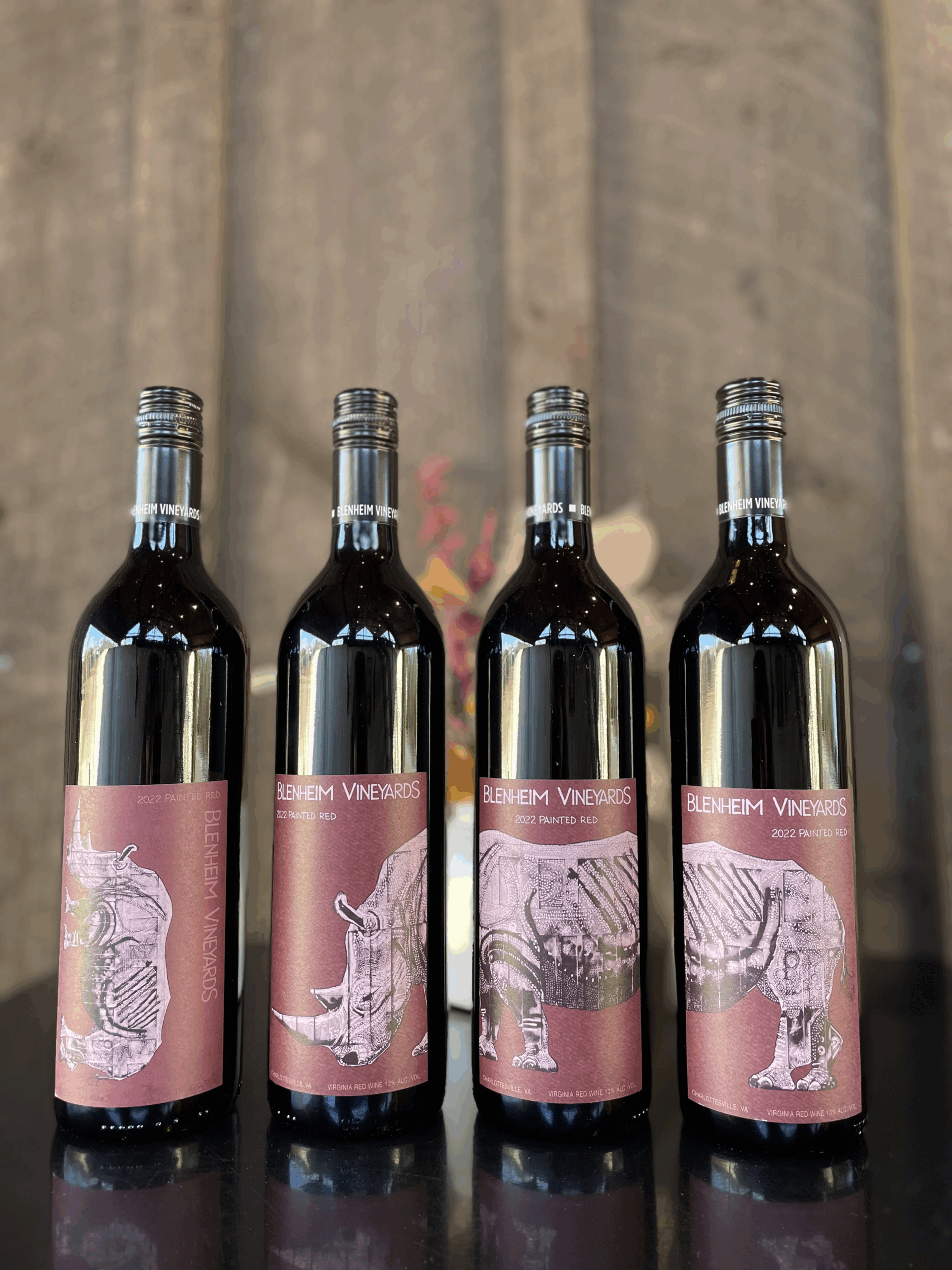 Four bottles of Blenheim Vineyards 2022 Painted Red wine displayed in a row on a reflective black surface. Each bottle features a unique burgundy label with a stylized rhinoceros illustration in a sketch or etching style. The background is softly blurred, with hints of wooden panels and floral decor, emphasizing the wine's artistic branding.