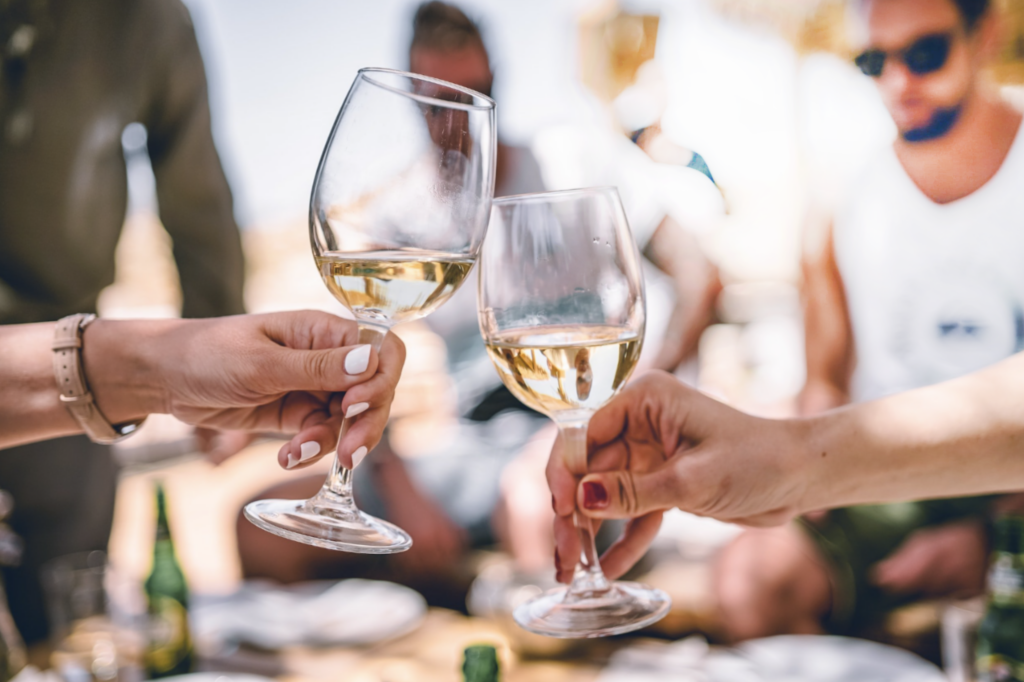 Two people toast with glasses of white wine in an outdoor setting. The background shows a blurred group of casually dressed individuals enjoying the sun, with bright lighting and a relaxed, summery atmosphere.