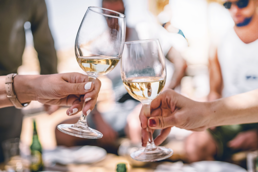 Two people toast with glasses of white wine in an outdoor setting. The background shows a blurred group of casually dressed individuals enjoying the sun, with bright lighting and a relaxed, summery atmosphere.