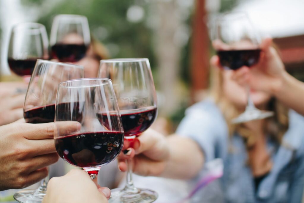 A group of people toasting with glasses of red wine outdoors. The image captures several hands holding glasses in a celebratory gesture, with a blurred background of greenery and soft natural light, suggesting a relaxed social gathering.