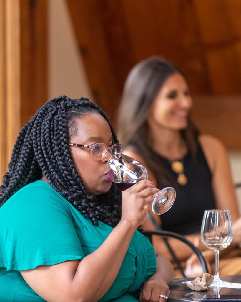 A woman in a green top with braided hair and glasses takes a sip of red wine from a glass during an indoor wine tasting event. Another woman in a black sleeveless top smiles in the blurred background, seated at a table with multiple wine glasses. The setting has warm wooden interiors.